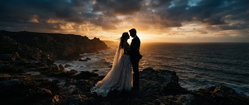 Intimate wedding portrait, a couple silhouetted against a moody sunset over a rugged coastline, cinematic wide aspect ratio, warm golden highlights.